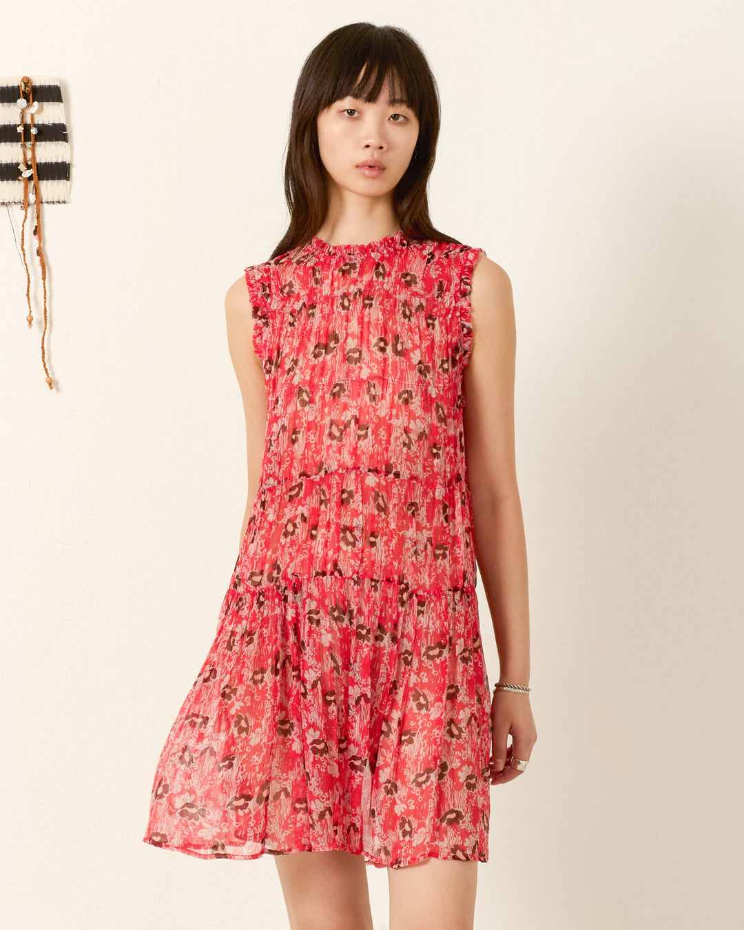 A woman wears the Jule Rougewood Dress—sleeveless and red with a 70s floral motif—in an indoor setting against a light wall, with a small decorative piece visible on the left.
