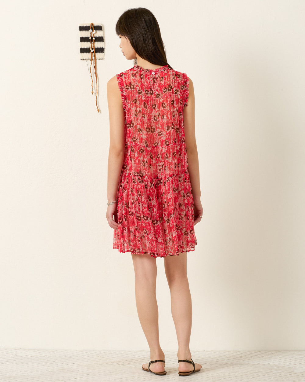 A person wearing the Jule Rougewood Dress—a sleeveless, red 70s-inspired floral dress—and sandals stands facing a white wall with a small woven hanging.