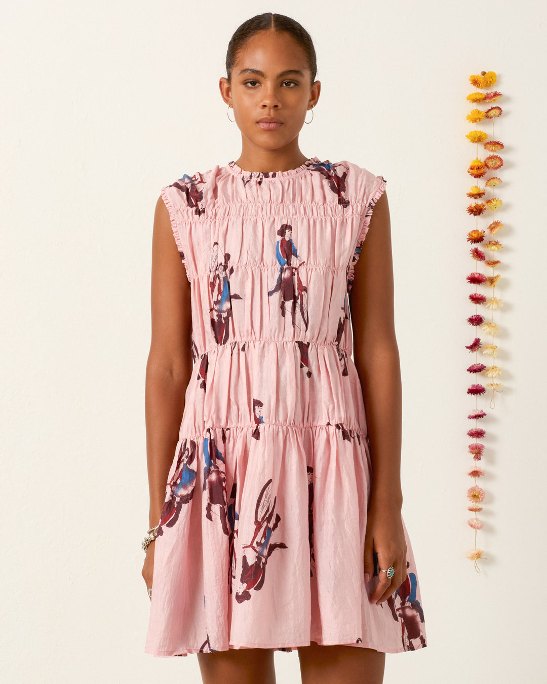 A woman wears the Jule Parisian Pedal Dress—sleeveless, light pink, hand-loomed silk with a gathered bodice and horse-and-rider print—standing by a white wall and a hanging strand of dried flowers.
