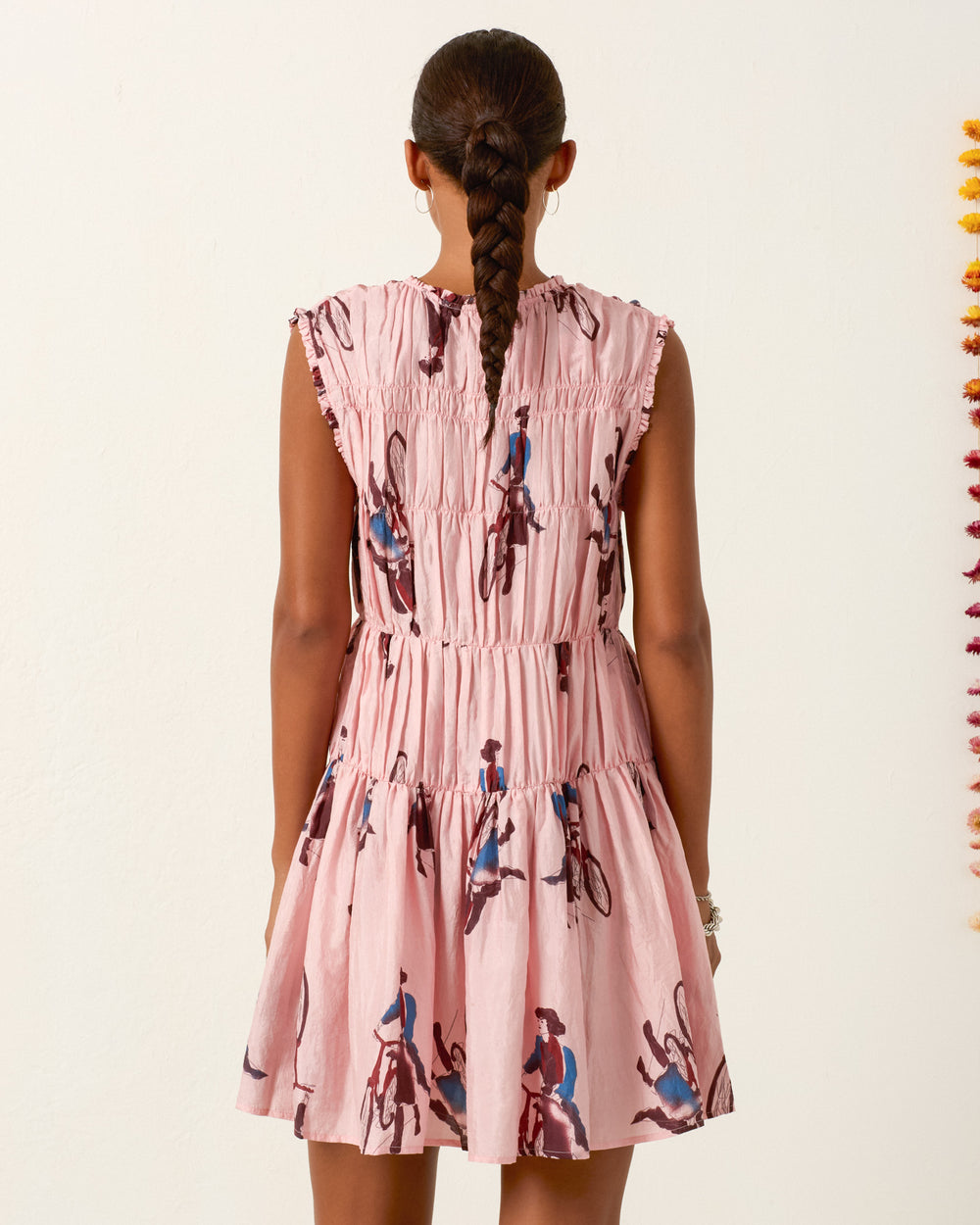A woman with a braided ponytail faces away, wearing the Jule Parisian Pedal Dress—a sleeveless pink dress with a gathered waist and vintage cyclist prints.