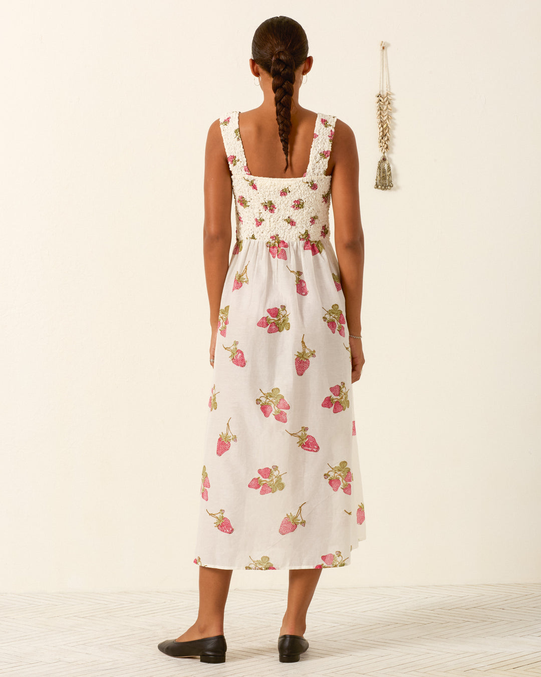 A woman with braided hair stands facing away, wearing the Kay Strawberry Dot Dress—a sleeveless white cotton-silk maxi dress with a crochet top and strawberry print skirt—paired with black flats. A tassel wall hanging is on the right.