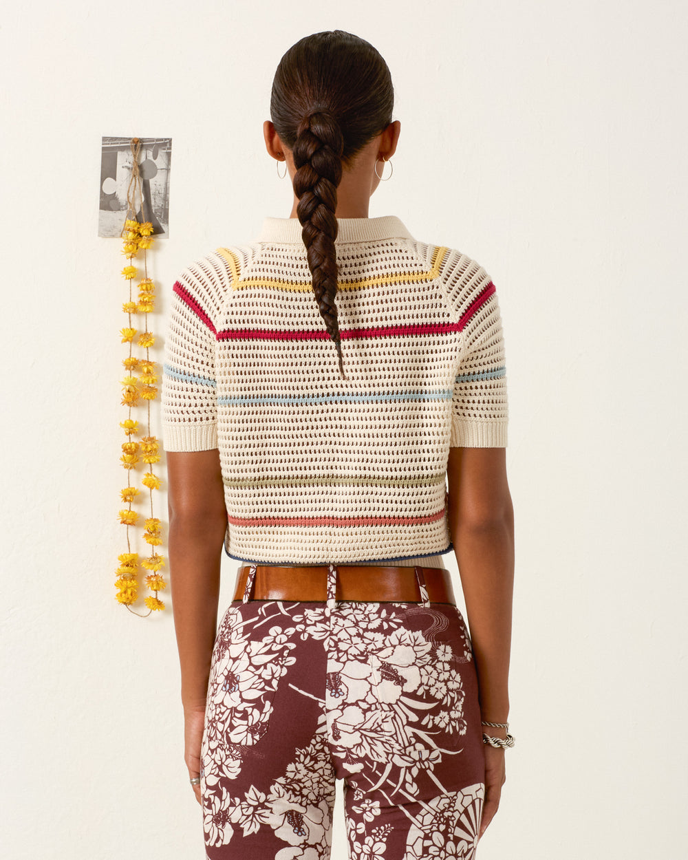 A woman faces a white wall, displaying her braided hair while wearing the Lessi Ivory Carnival Top and brown floral pants. A string of yellow flowers and a photo are pinned to the wall behind her.
