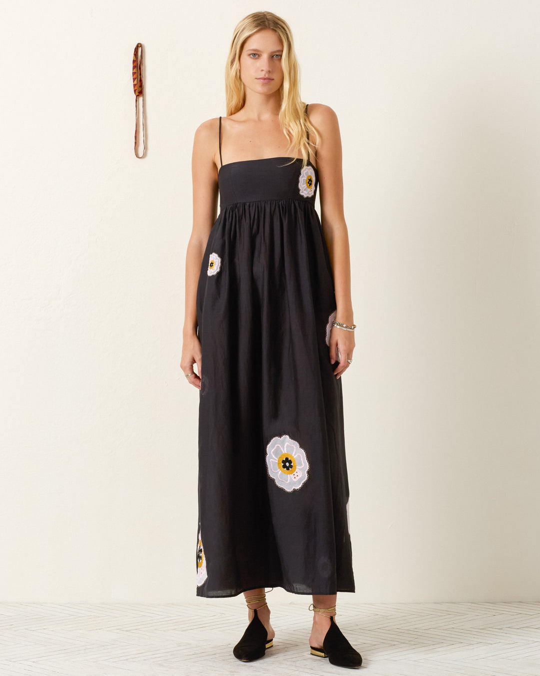 A woman stands indoors wearing the Lilia Pressed Daisy Dress, a long black empire-waist dress with thin straps and floral designs, paired with black flats and a bracelet. A wall decoration is visible behind her.
