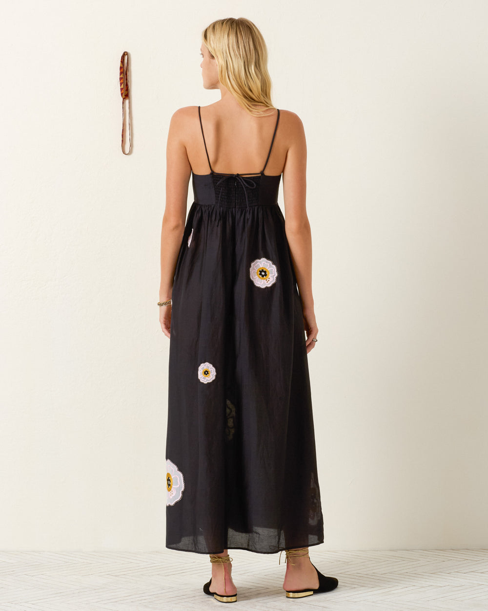A woman stands with her back to the camera in a minimal indoor setting, wearing the Lilia Pressed Daisy Dress—a long black maxi dress with thin straps and white daisy floral designs.