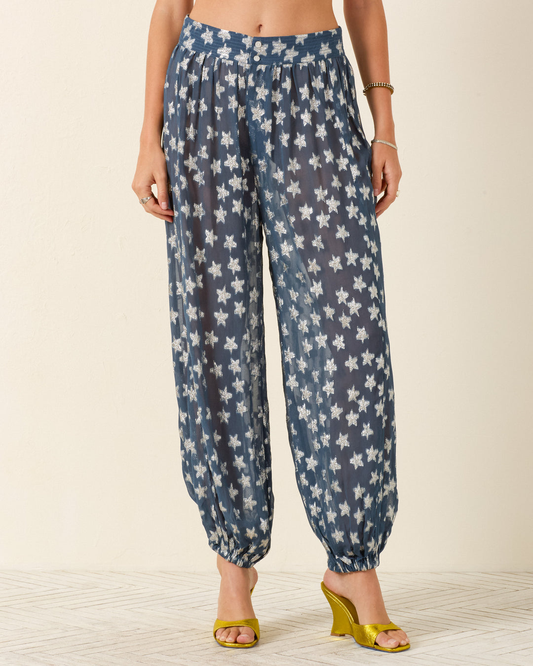 A person wears the Margot Starlite Pant—blue harem-style viscose-chiffon with metallic silver stars—and yellow open-toe heels, standing against a plain background.