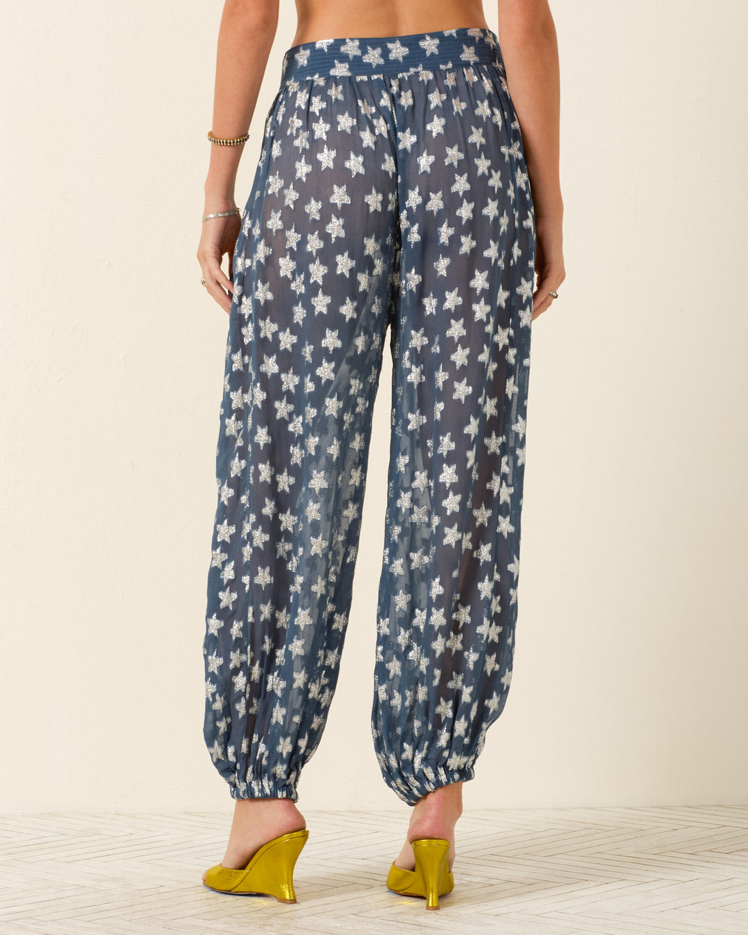 A person shown from the back wears the Margot Starlite Pant in blue viscose-chiffon with a white floral pattern, paired with yellow heeled shoes.