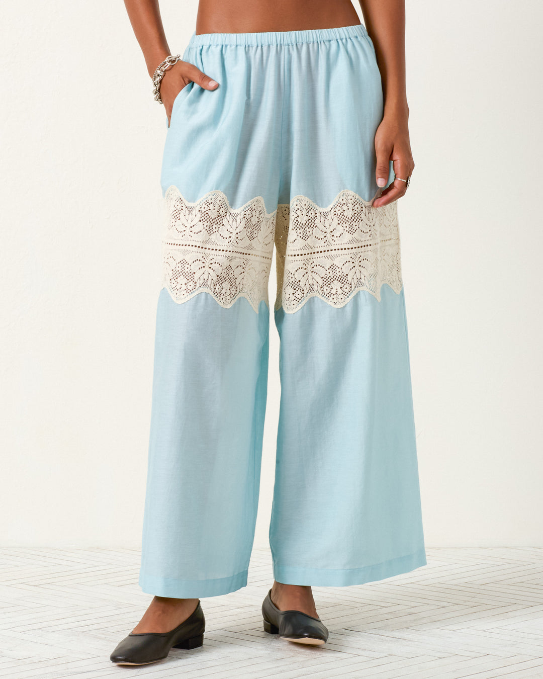 A person wearing Margie Sky Prima Lace Pant in light blue, featuring a cotton-silk blend and cream lace panel across the thighs, paired with black flats and a silver bracelet.