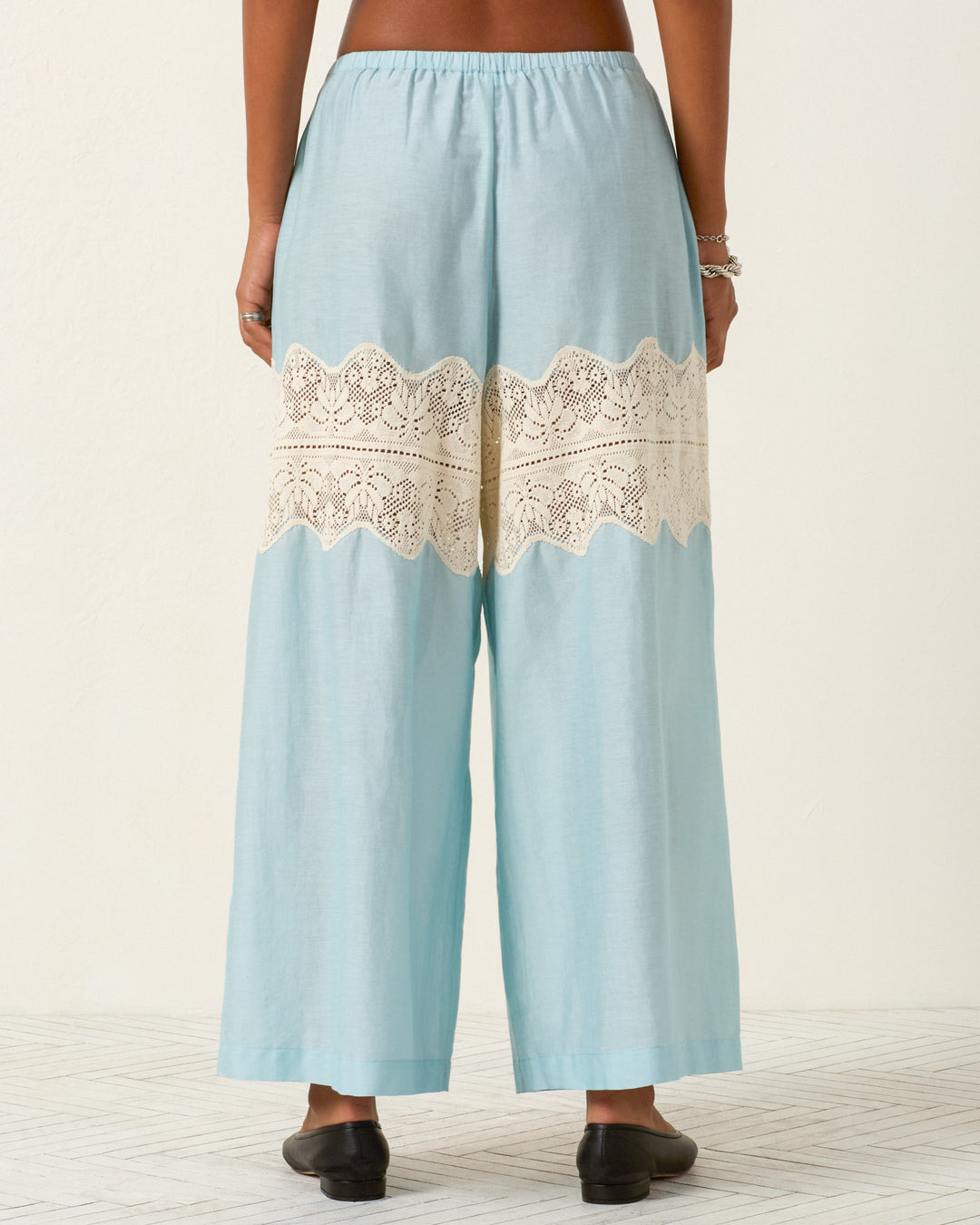 A person wearing Margie Sky Prima Lace Pant—light blue wide-leg pants with a cream cotton lace panel across the back—stands on a light patterned floor.