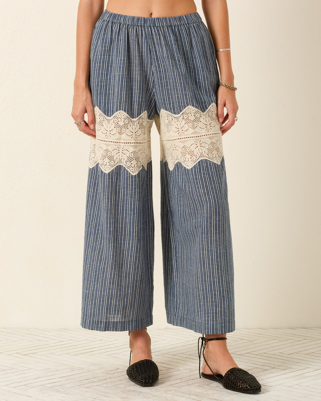 A person wearing the Margie Prima Micro Plaid Pant, featuring wide-leg khadi cotton in a blue check pattern with a cream lace panel across the thighs, paired with black woven flats.