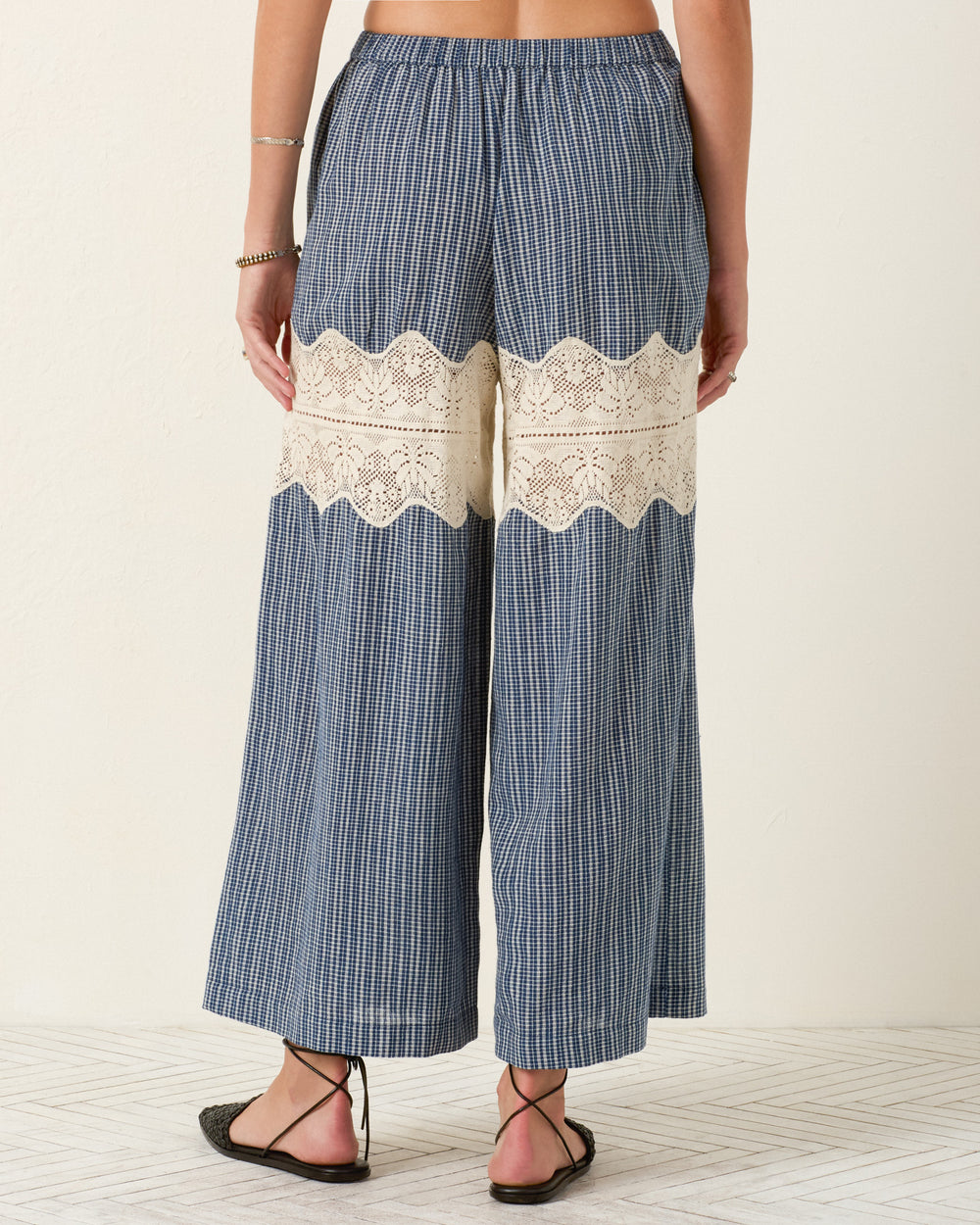 A person wears the Margie Prima Micro Plaid Pant—wide-leg blue plaid Khadi cotton pants with a cream lace band across the back thighs—standing on a light patterned floor.