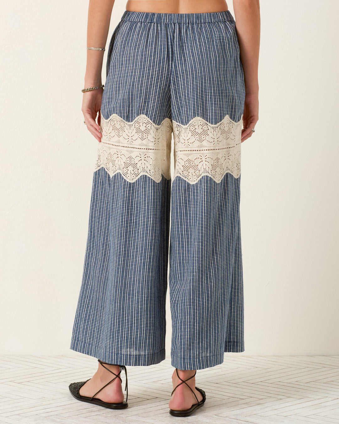 A person wears the Margie Prima Micro Plaid Pant—wide-leg blue plaid Khadi cotton pants with a cream lace band across the back thighs—standing on a light patterned floor.