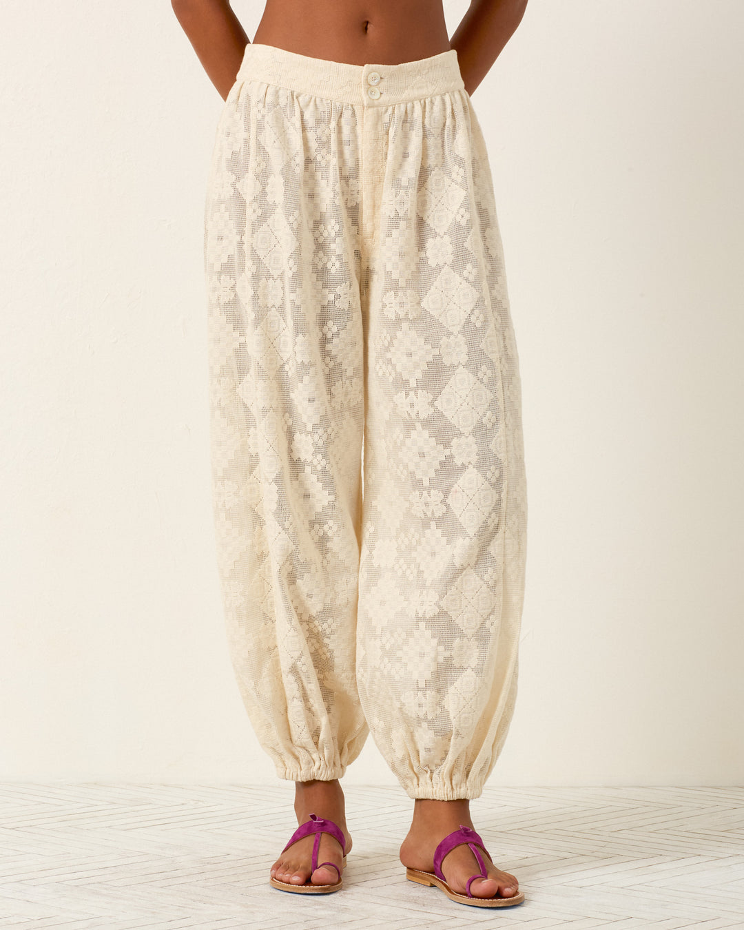 A person wears the Margot Ecru Tetris Lace Pant—loose, cream-colored with a geometric motif and elastic cuffs—paired with magenta sandals, standing against a plain light background.