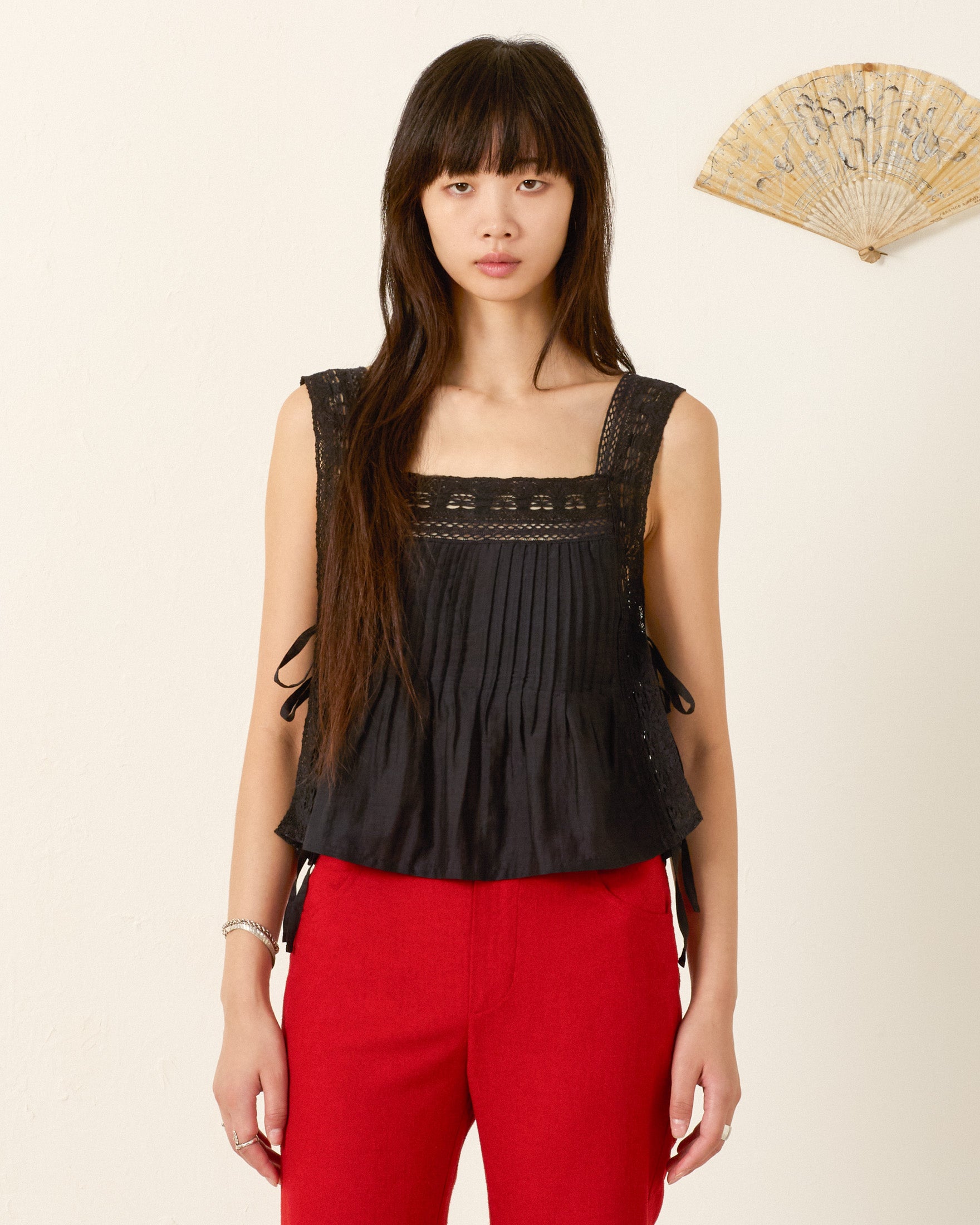 A woman stands against a light wall with a decorative fan, wearing the Mimi Onyx Prima Lace Top—a black sleeveless cotton-silk blend lace top with pleats—and red pants.