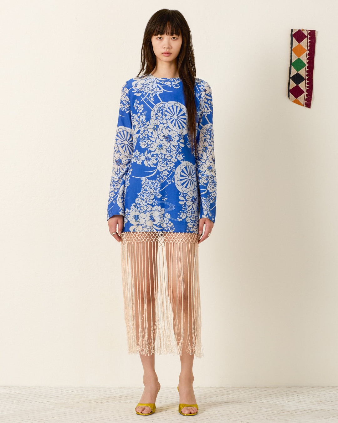 A woman stands against a plain wall, wearing the Narene Royal Spoke Dress in blue floral with beige fringe at the hem and yellow shoes. To her right, a small geometric textile decor with a hanaguruma motif hangs on the wall.