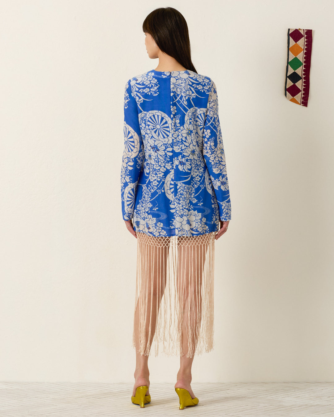 A woman faces away, wearing the Narene Royal Spoke Dress—a blue and white floral mini dress with beige fringe detail—and yellow heels, beside a colorful wall hanging on a plain background.