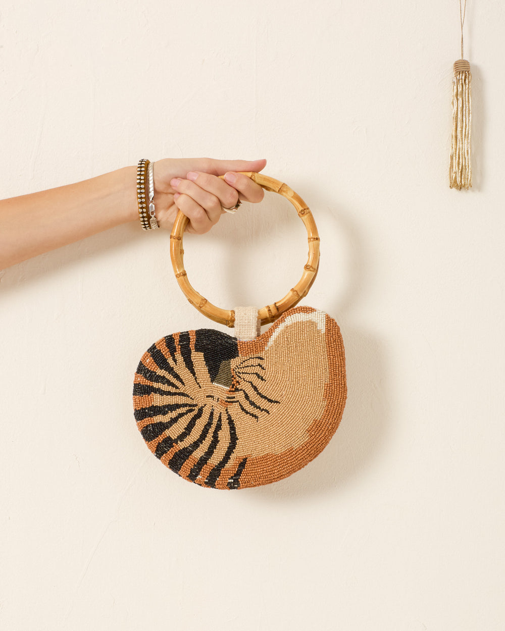 A hand displays the Nautilus Sand Bag, a chic, hand-beaded accessory with a bamboo handle and nautilus shell motif in tan, black, and brown against a plain white wall.