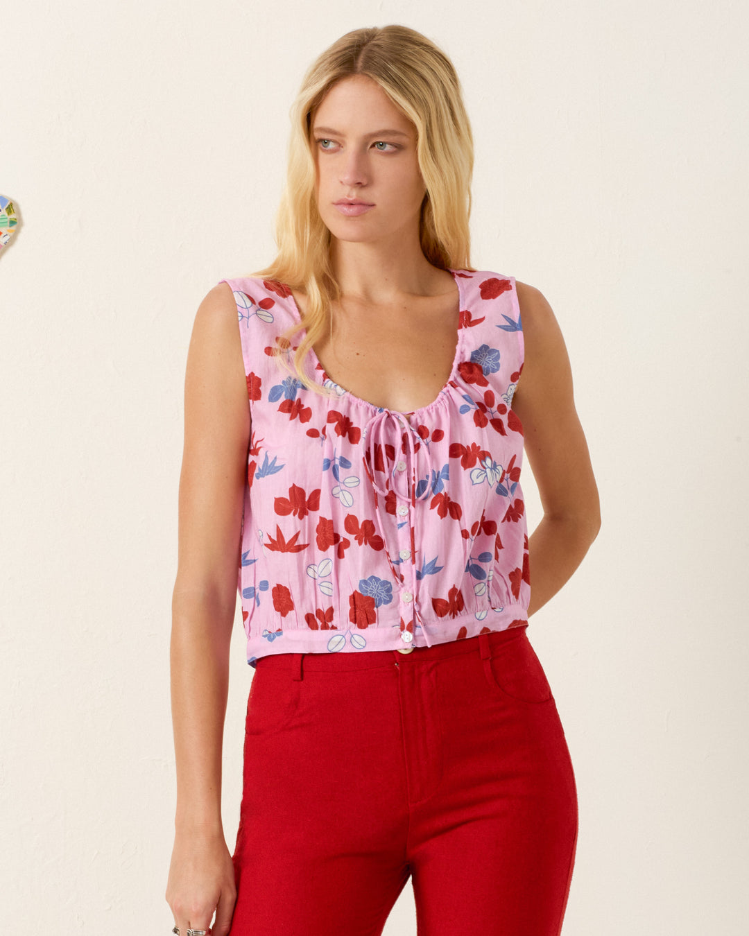 A woman models the Nova Tutu Bloom Shirt, a sleeveless summer top featuring a pink, red, and blue floral pattern, paired with red high-waisted pants against a plain light-colored wall.