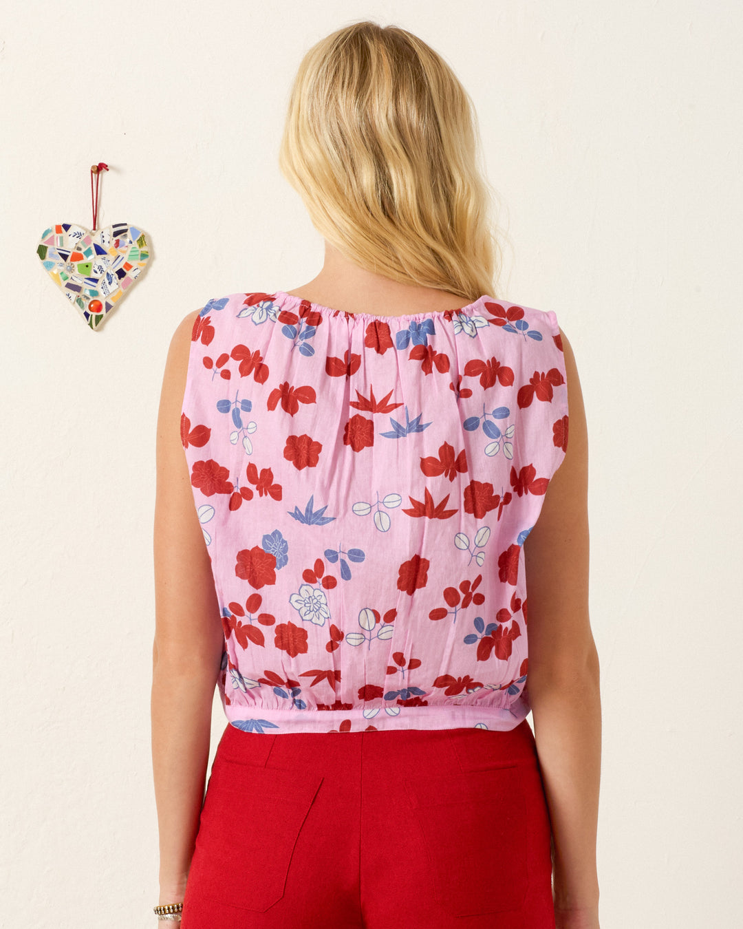 A person with blonde hair faces away, wearing the Nova Tutu Bloom Shirt, a pink kimono-inspired blouse with red floral patterns and red pants. A mosaic heart decor hangs on the wall to the left.