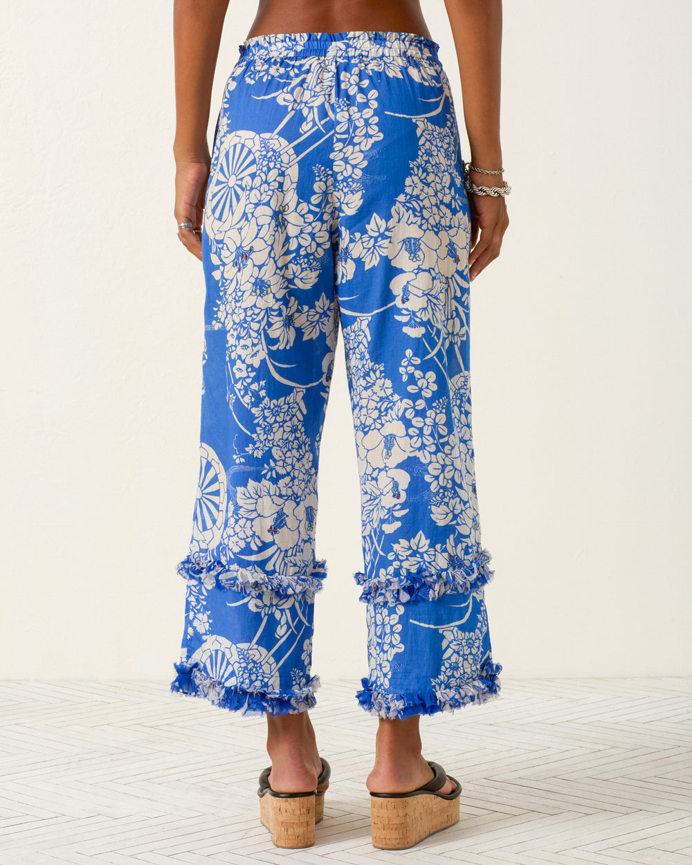 Person wearing the Otto Royal Spoke Pant, featuring a blue and white floral print in cotton voile, pajama-style silhouette, and fringed hems, viewed from the back while standing on a light-colored floor.