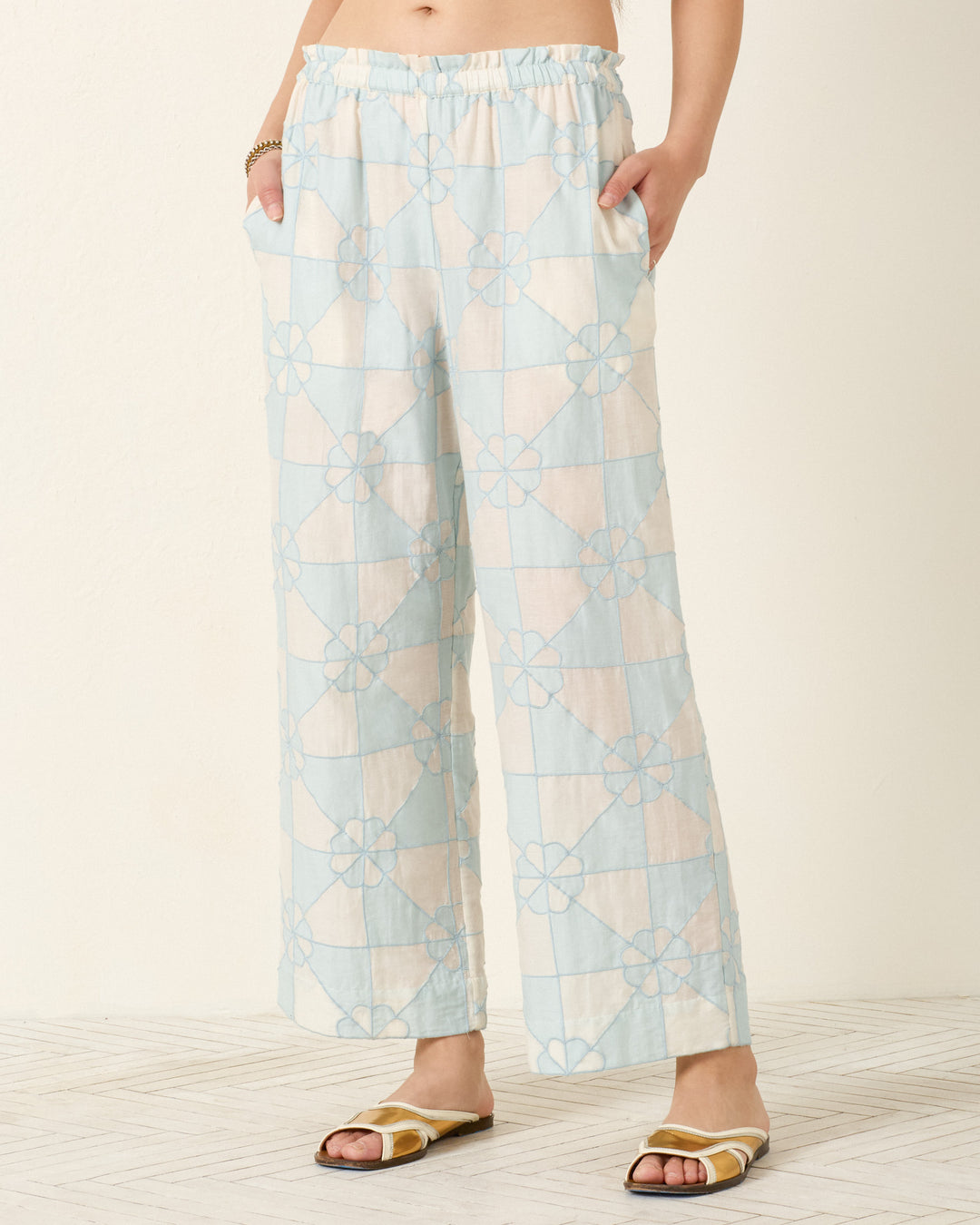 Someone wearing the Otto Sky Jubilee Pant—light blue and white patchwork with a floral geometric pattern and pajama-style silhouette—stands on a light-colored floor, hands in pockets.