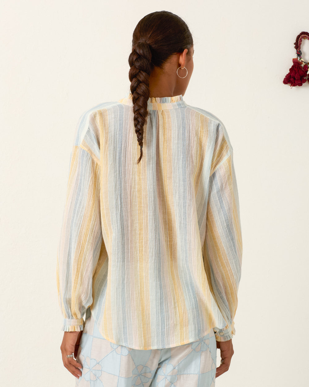 A person with a braided hairstyle is wearing the Poet Sunwash Stripe Blouse, a loose, long-sleeved pastel striped shirt, and light blue patterned pants, standing with their back to the camera.