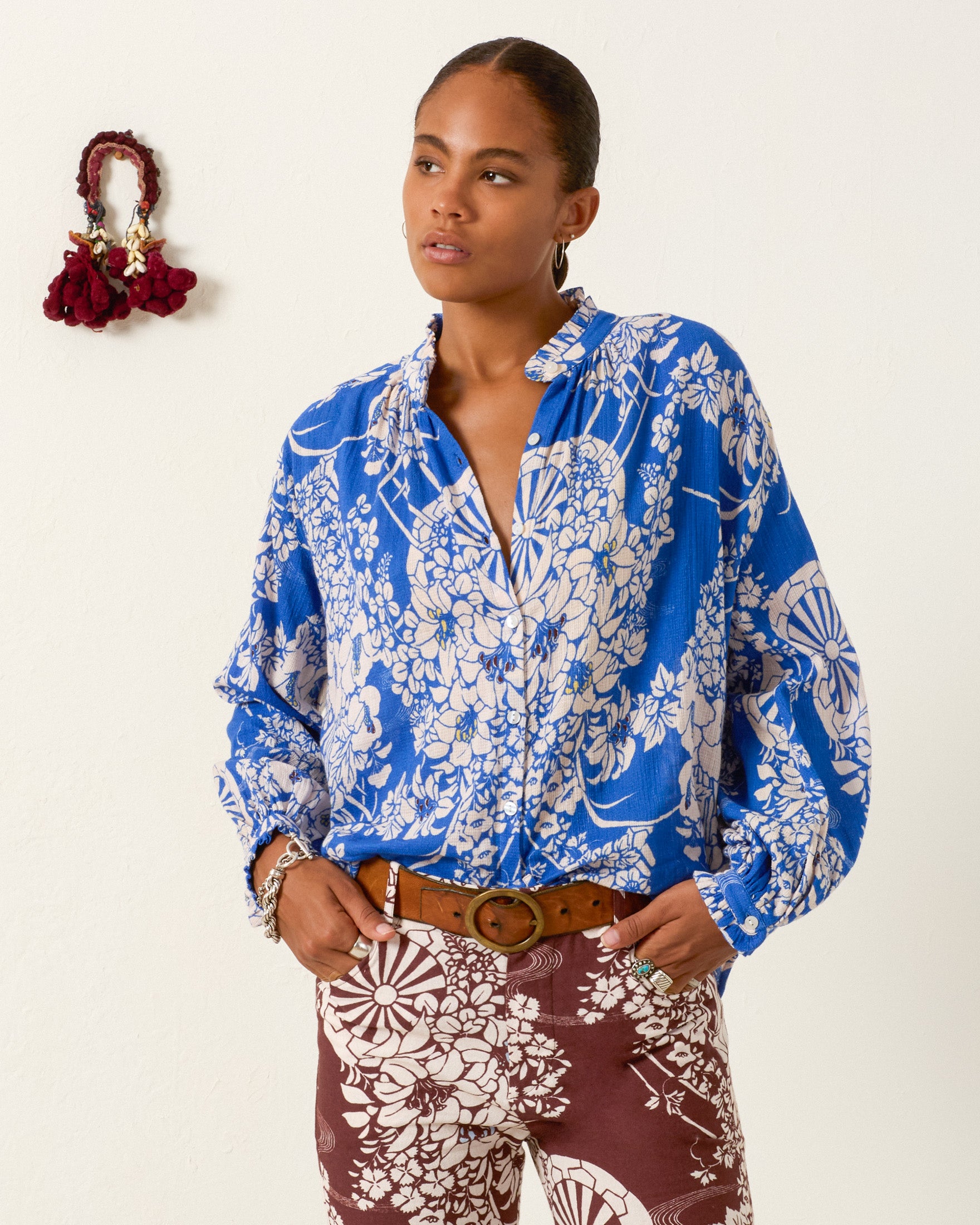 A person stands against a light wall wearing the Poet Royal Spoke Blouse and brown floral pants, one hand in their pocket, with a small decorative item mounted on the wall.