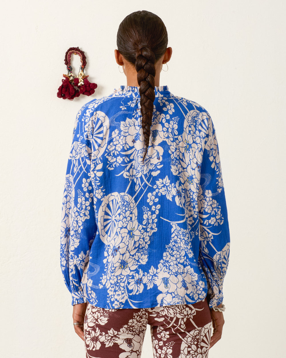 A person with a braided ponytail stands with their back to the camera near a white wall, wearing the Poet Royal Spoke Blouse in blue and white floral print paired with brown floral pants and a red hair accessory.