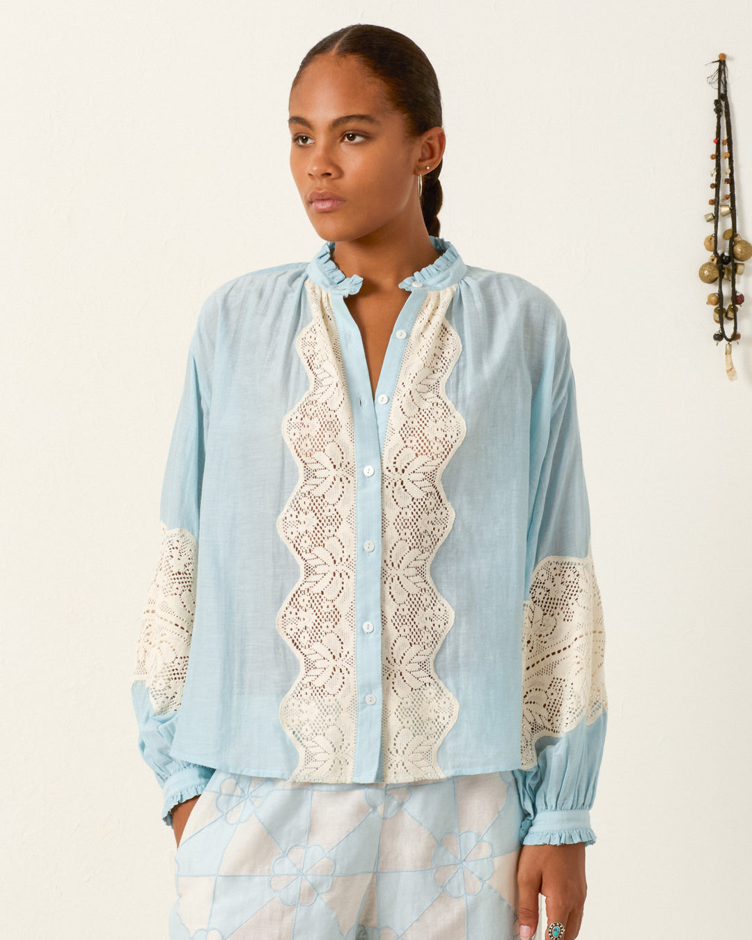 A person wears the Poet Sky Prima Lace Blouse, featuring vintage-inspired cotton lace panels, paired with patterned light blue pants and stands against a white wall with a beaded wall hanging in the background.