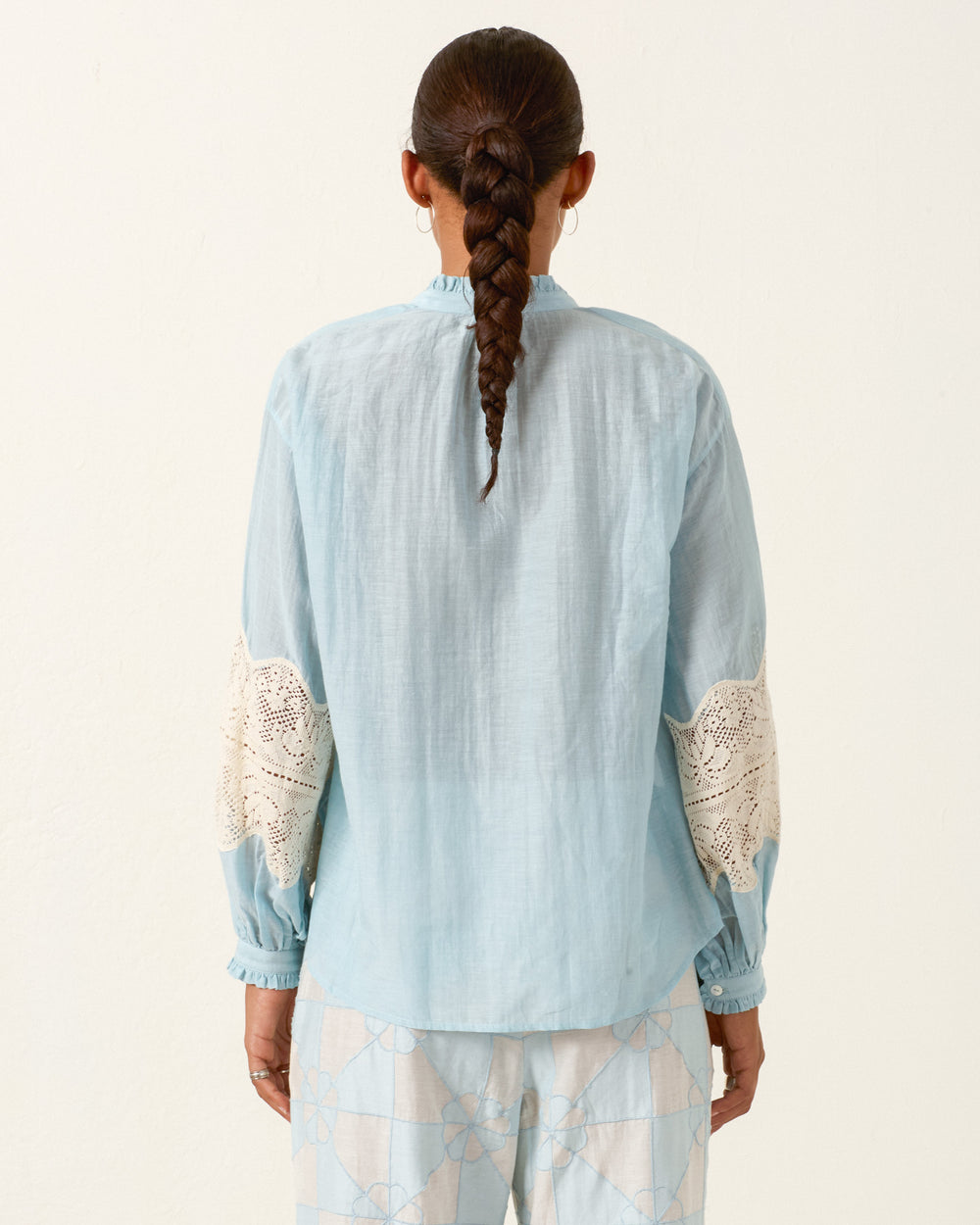 Person shown from the back with braided hair, wearing the Poet Sky Prima Lace Blouse—a vintage-inspired light blue cotton-silk shirt with lace elbow patches—and patterned pants.