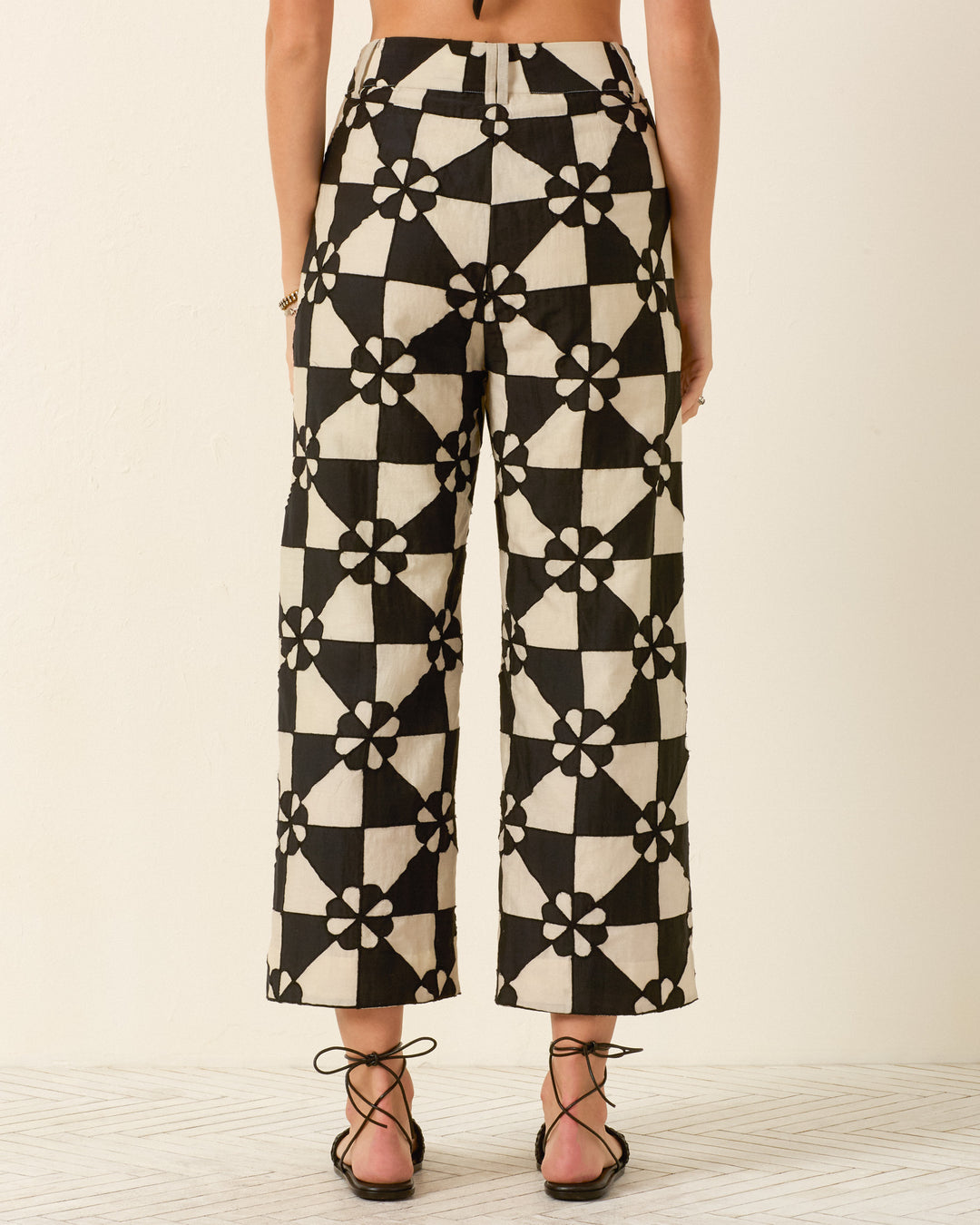 Someone wearing the Scout Onyx Jubilee Pant—high-waisted, wide-leg pants with black and white geometric and floral patchwork—paired with black strappy sandals, viewed from the back.