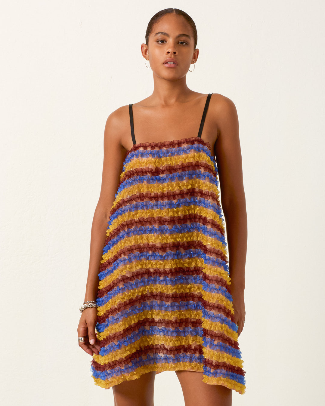 A person wearing the Soleil Jester Vale Dress—a sleeveless, multicolored striped trapeze dress with textured horizontal bands—stands against a plain light background.