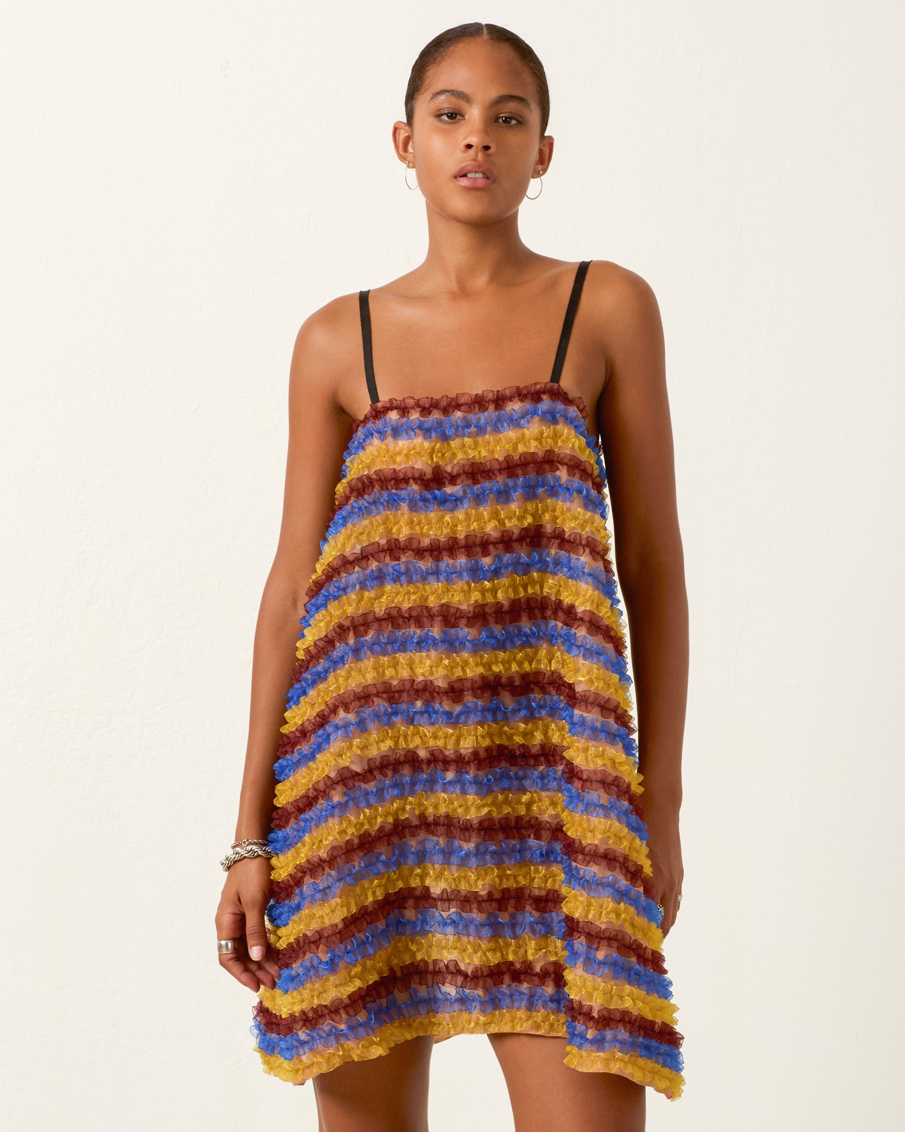 A person wearing the Soleil Jester Vale Dress—a sleeveless, multicolored striped trapeze dress with textured horizontal bands—stands against a plain light background.