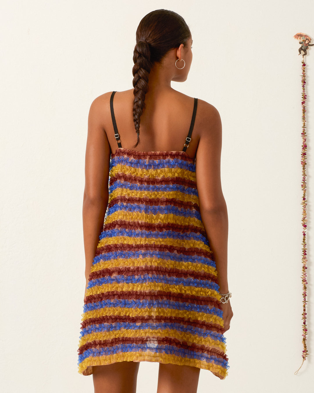 A woman with a braided ponytail models the Soleil Jester Vale Dress, a sleeveless, striped trapeze style in brown, gold, and blue hues. She stands by a plain wall with a hanging string of small objects on the right.
