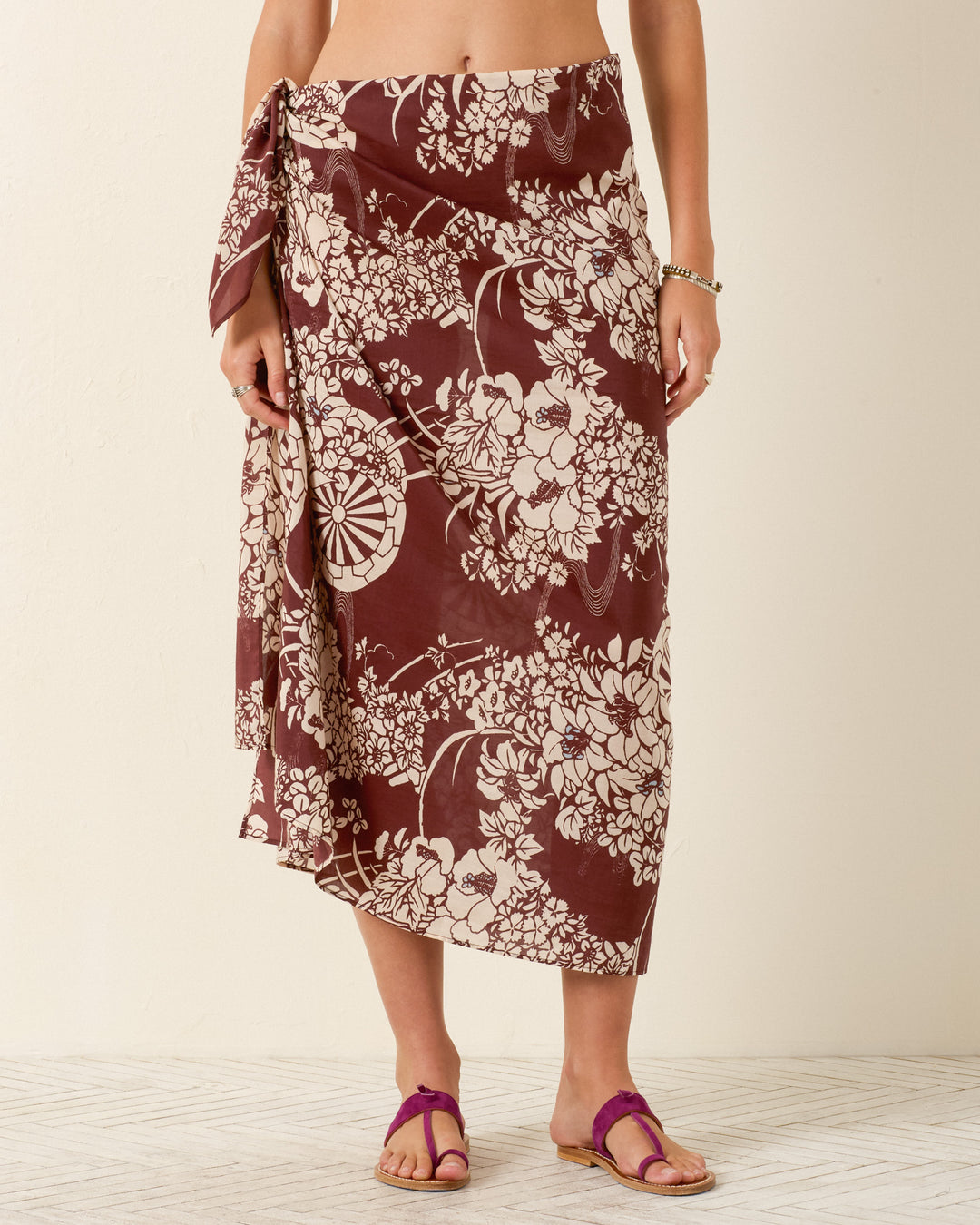 A person stands barefoot, wearing the Soraya Auburn Spoke Sarong—maroon and white with a hanaguruma motif—and purple sandals on a light textured floor.