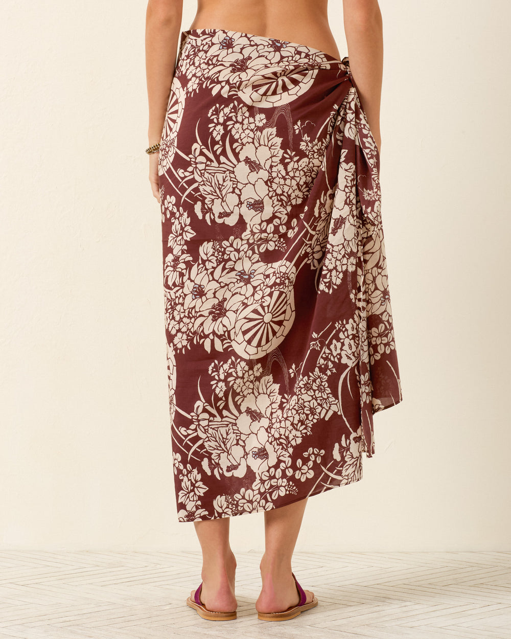 A person stands barefoot, shown from the back against a neutral background, wearing the Soraya Auburn Spoke Sarong—a brown and white wrap skirt inspired by artisan textile traditions and tied at the waist.