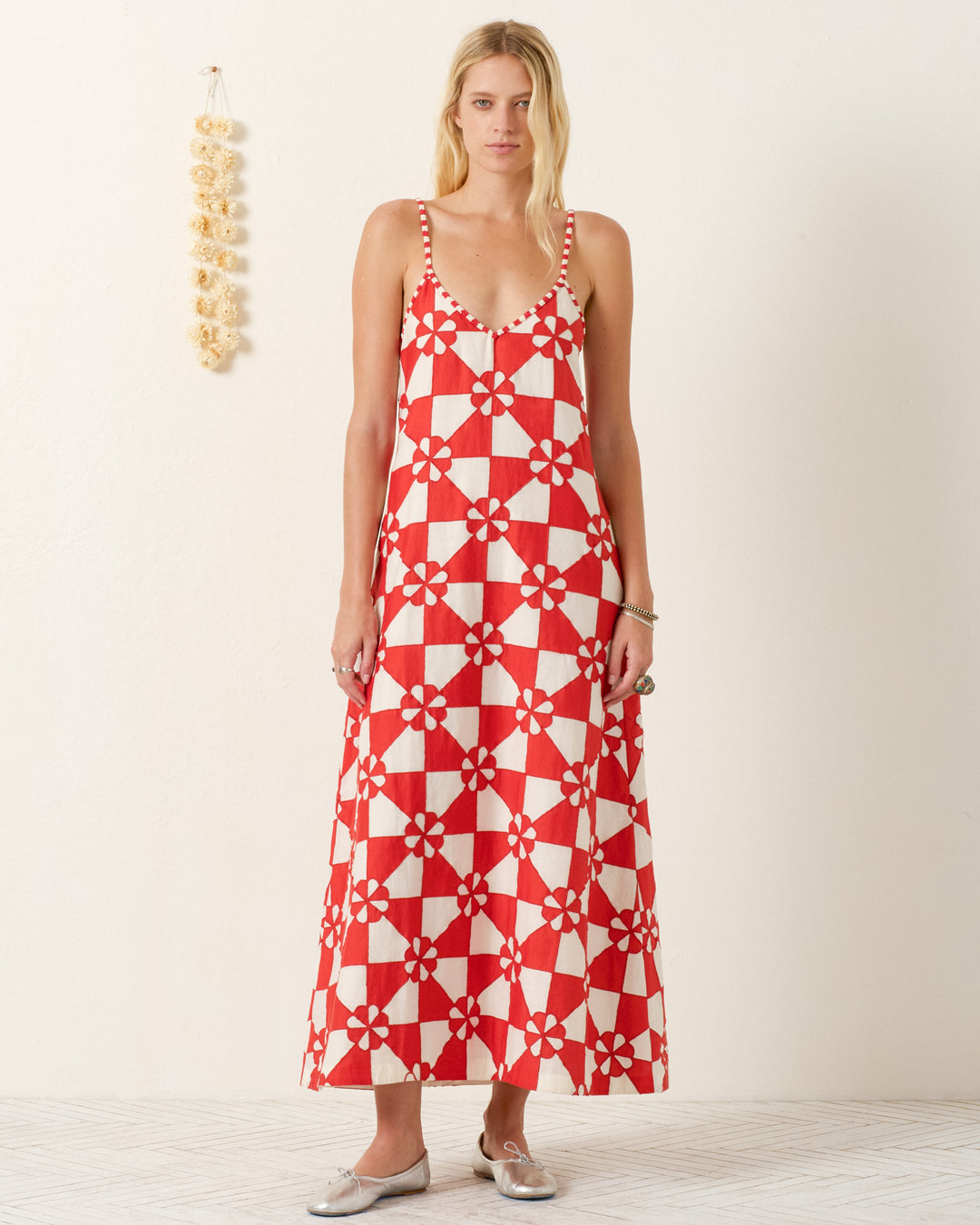 Woman wearing the Spice Ibis Jubilee Dress, a long sleeveless patchwork slip dress with a red and white geometric floral pattern, paired with silver flat shoes, standing against a plain light wall.
