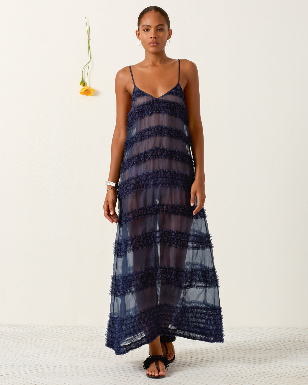 A woman stands indoors wearing the Spice Market Jester Eve Dress, a navy blue sheer slip with horizontal textured stripes, paired with black sandals and minimal jewelry; a yellow flower hangs on the wall behind her.