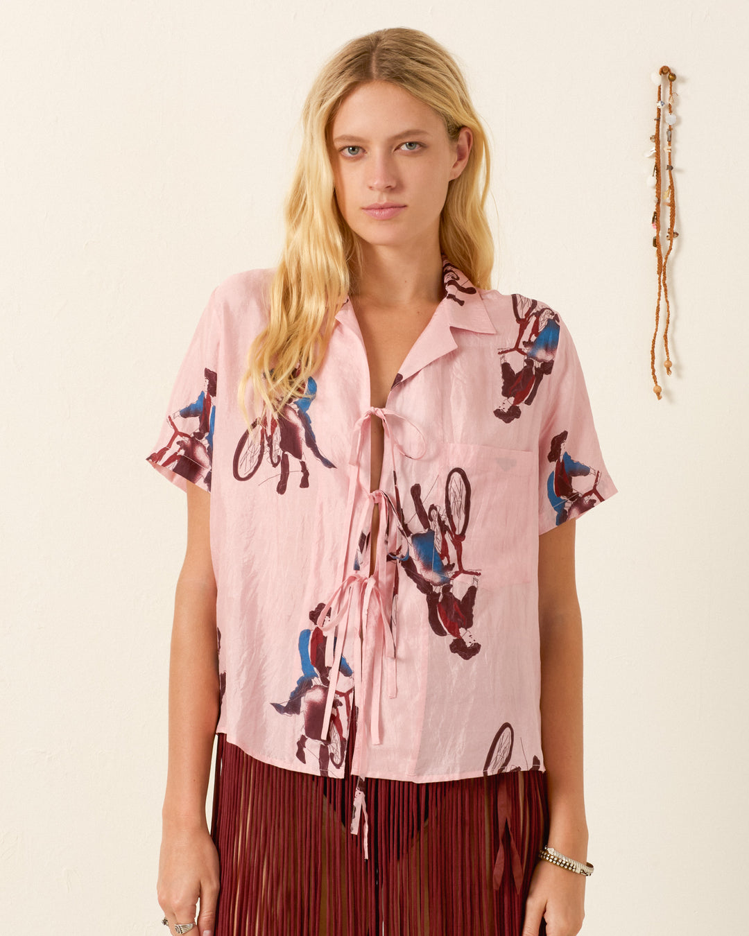 A woman wears the Styla Parisian Pedal Shirt—light pink, hand-loomed silk with a vintage cyclist motif and front ties—standing against a plain wall with a beaded decoration on the right.