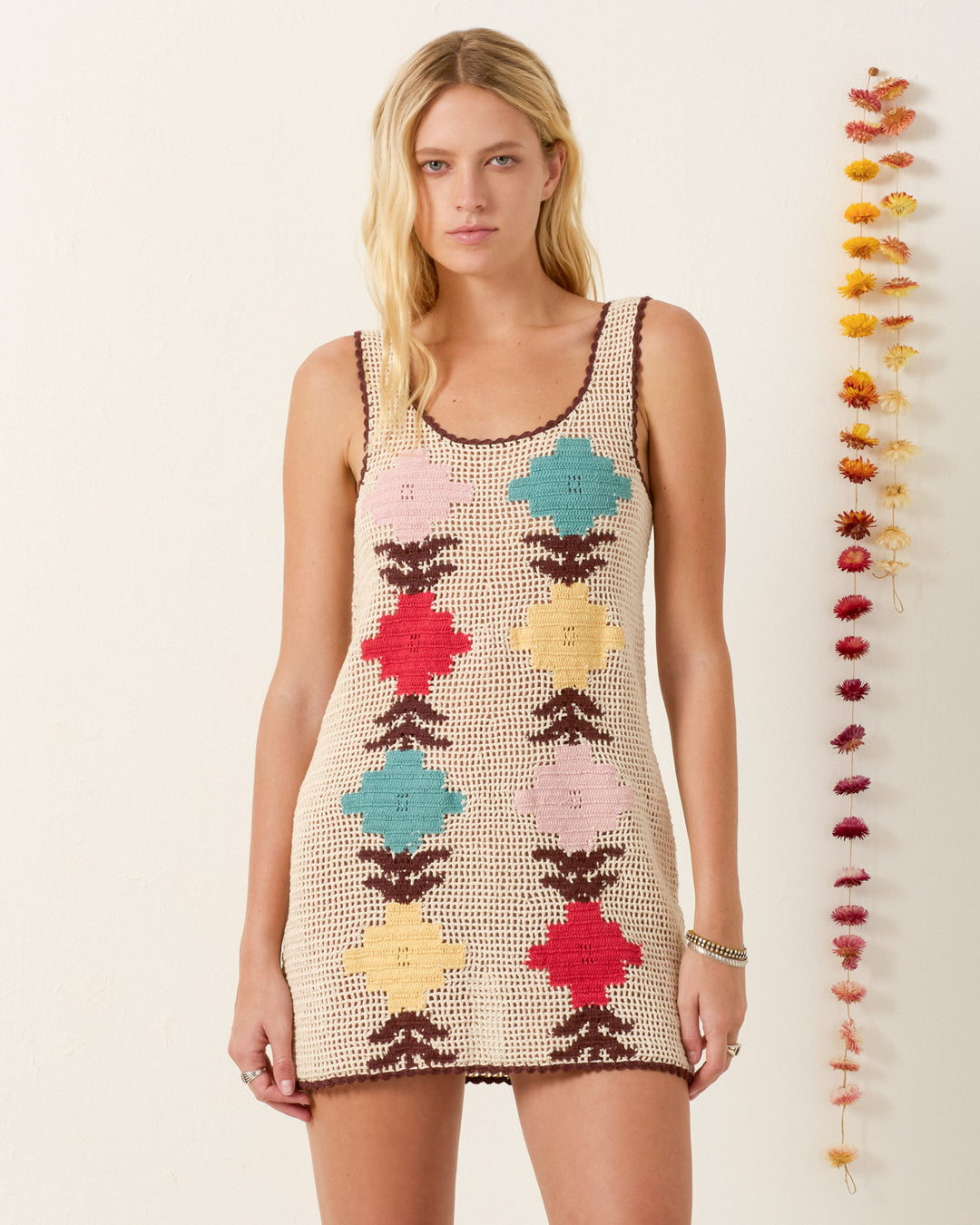 A woman stands by a white wall adorned with dried flowers, wearing the Tyler Daisy Chain Dress—a sleeveless, artisan-made crochet mini dress featuring colorful floral patterns.