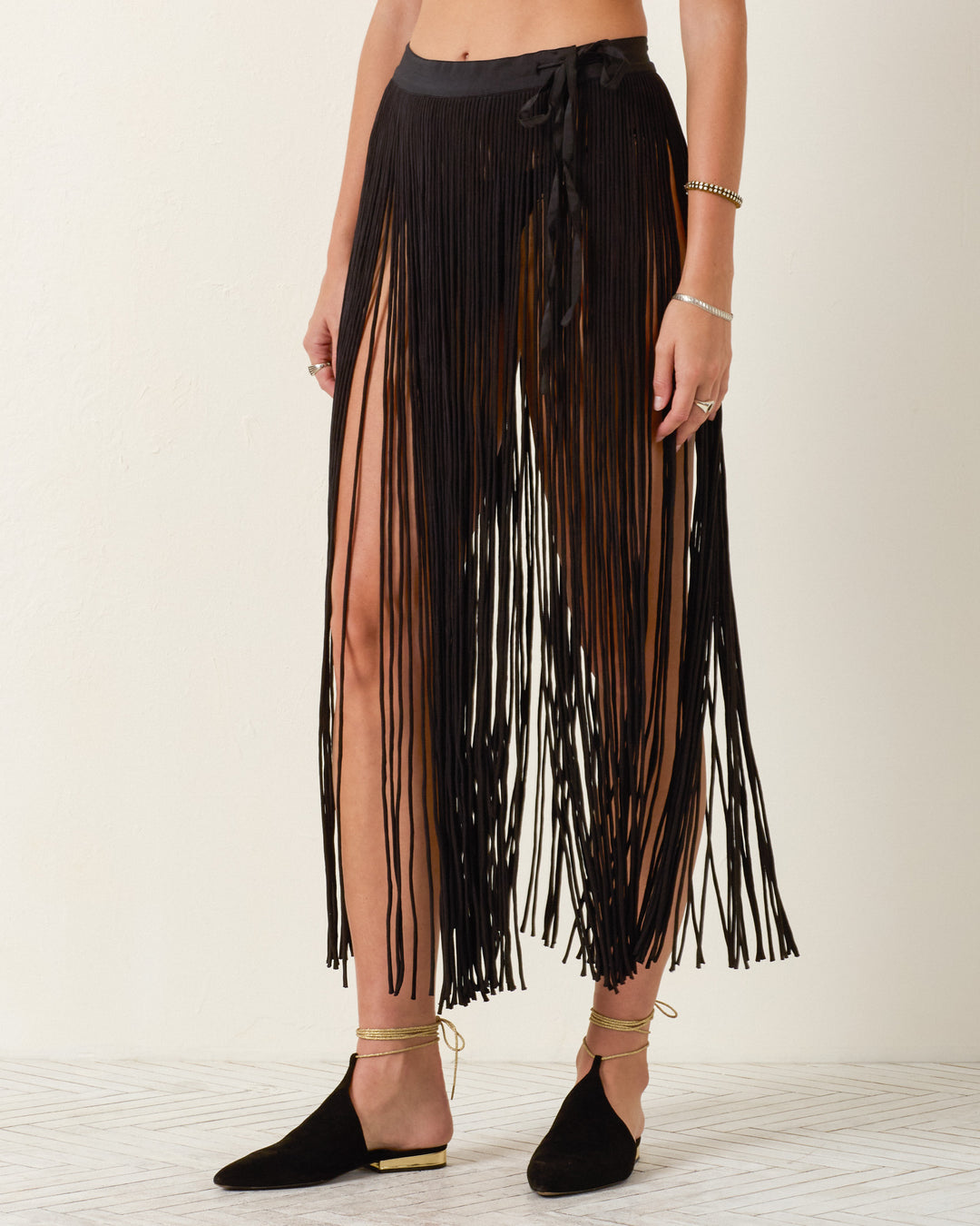 Wearing the Vanessa Onyx Fringe Skirt, paired with black flats and gold anklets, a person poses against a plain light background.