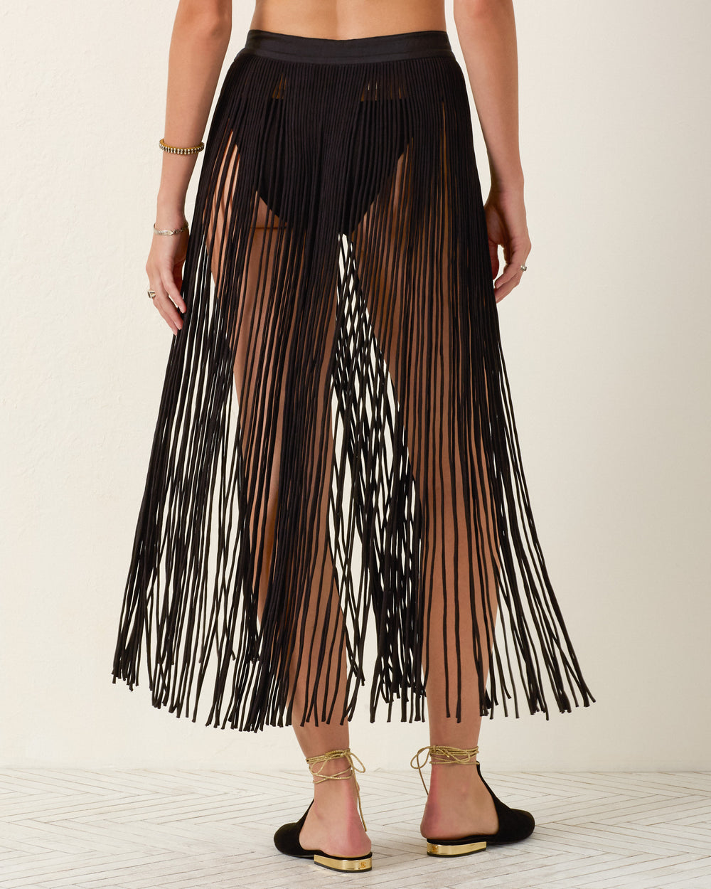A person wearing the Vanessa Onyx Fringe Skirt over a black bikini bottom and black shoes, standing against a light background.