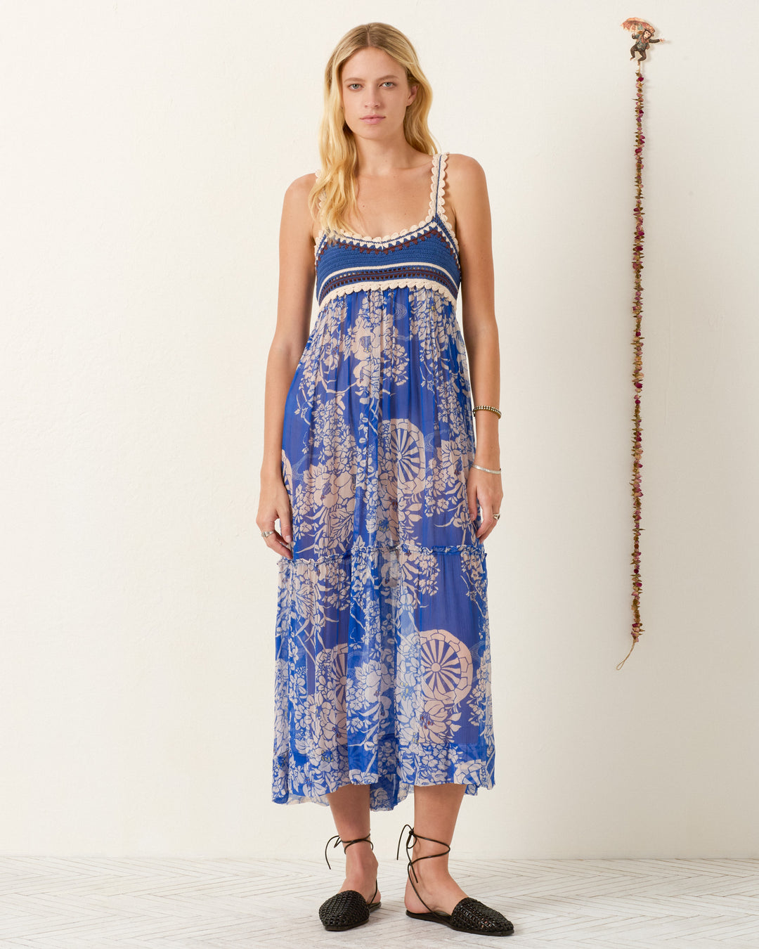 A woman stands against a plain wall with a hanging garland decoration, wearing the Victoria Royal Spoke Dress—an elegant empire-waist maxi dress featuring blue floral prints, sleeveless design, and crochet trim.