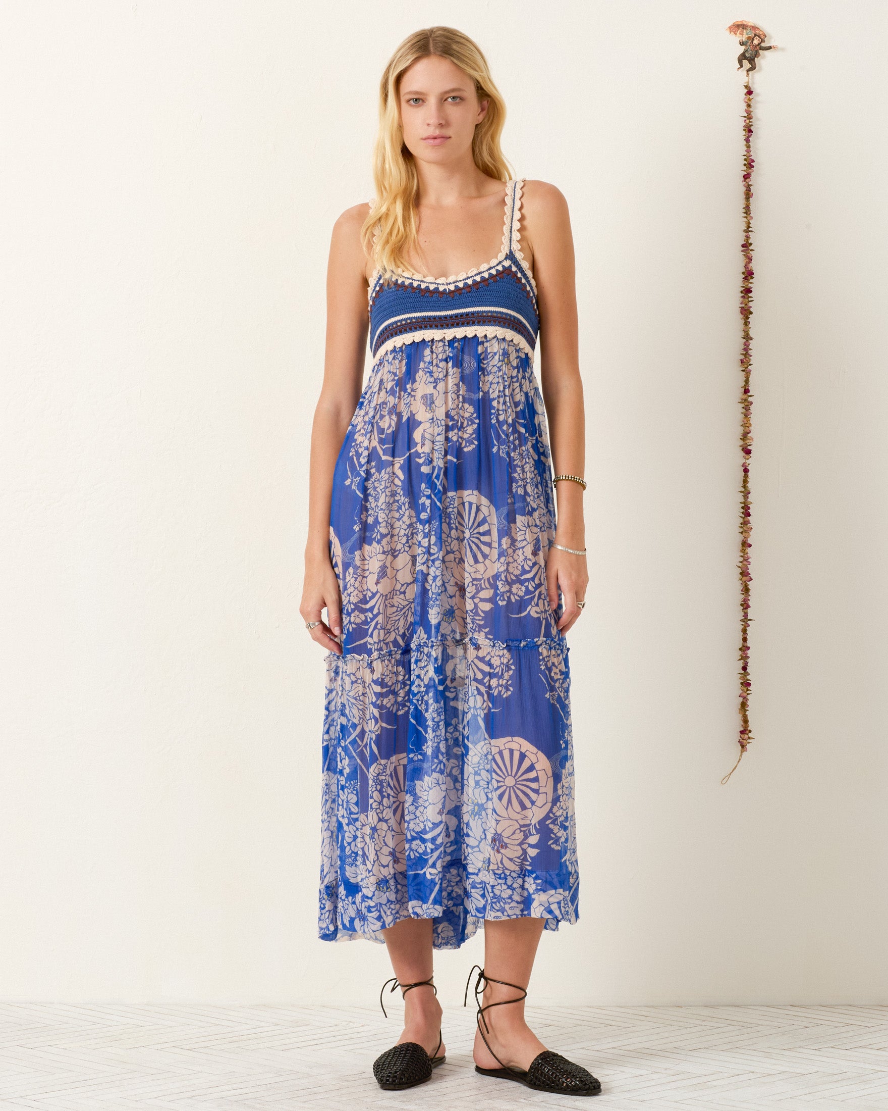 A woman stands against a plain wall with a hanging garland decoration, wearing the Victoria Royal Spoke Dress—an elegant empire-waist maxi dress featuring blue floral prints, sleeveless design, and crochet trim.