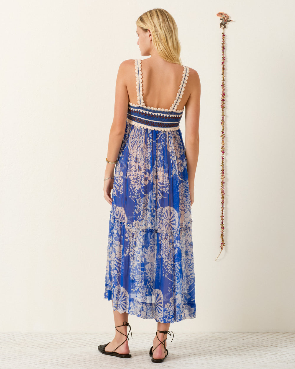 A woman stands with her back to the camera, wearing the Victoria Royal Spoke Dress—a blue and white handmade crochet dress with an empire-waist silhouette and delicate lace trim—paired with black sandals in a minimal room accented by a string of decorative items on the wall.