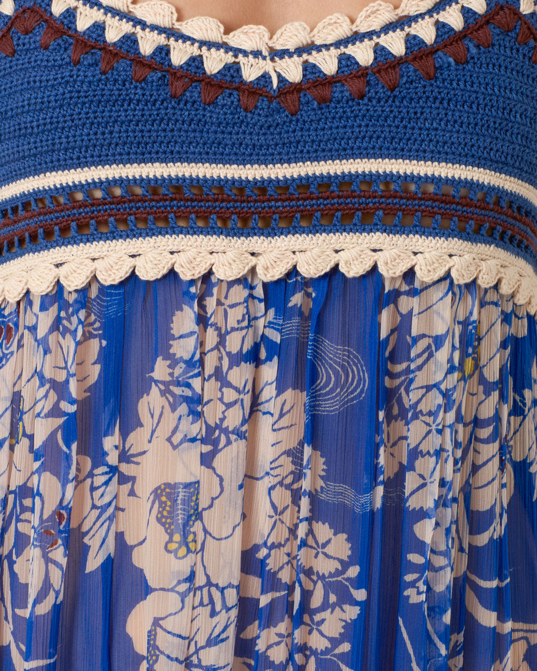 A close-up of the Victoria Royal Spoke Dress, showcasing its handmade crochet blue bodice with cream and brown trim, transitioning into an empire-waist silhouette with a pleated chiffon skirt featuring a blue and cream floral print.