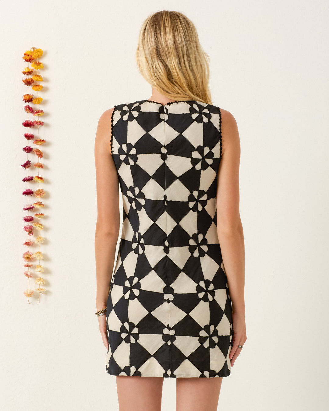 A woman with long blonde hair stands facing away, wearing the Vita Onyx Jubilee Dress; a vertical garland hangs on the wall to her left.