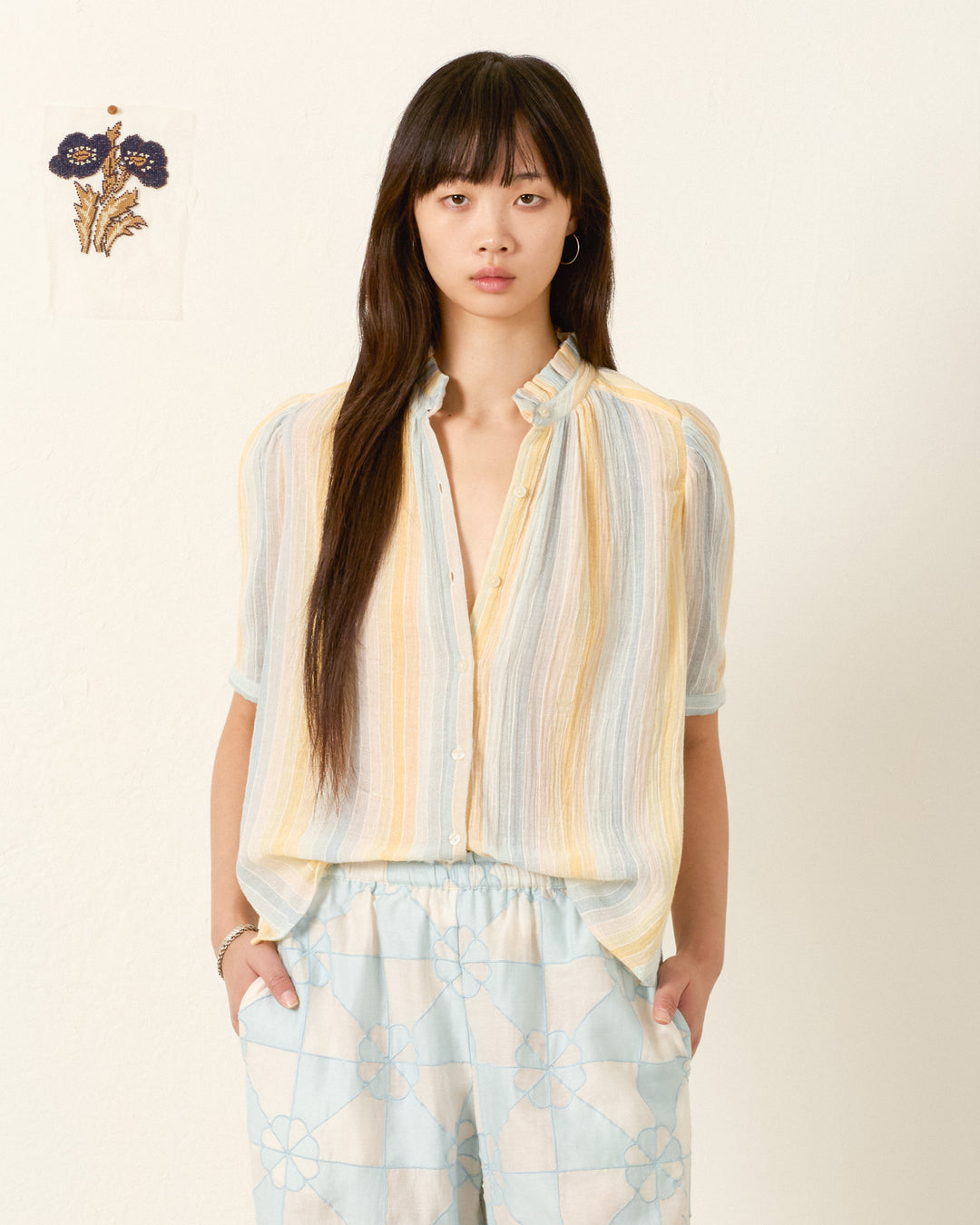 A woman with long dark hair wears the Winnie Sunwash Stripe Shirt, featuring a ruffled collar, and patterned light blue pants, standing against a plain wall with small floral artwork in the background.