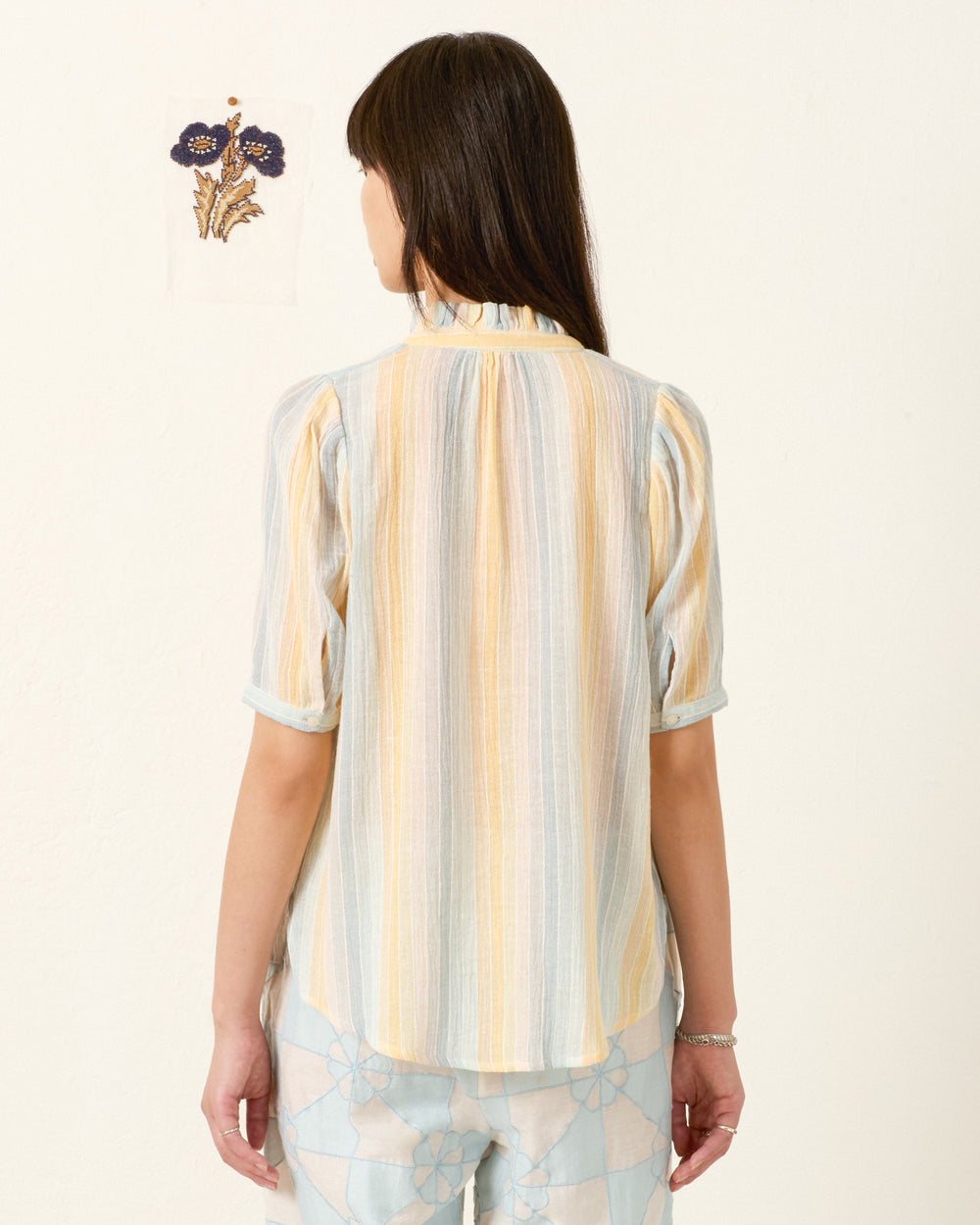 A woman faces away wearing the Winnie Sunwash Stripe Shirt, a light pastel-striped short-sleeve top in gauzy cotton, as an embroidered floral artwork hangs on the wall ahead.