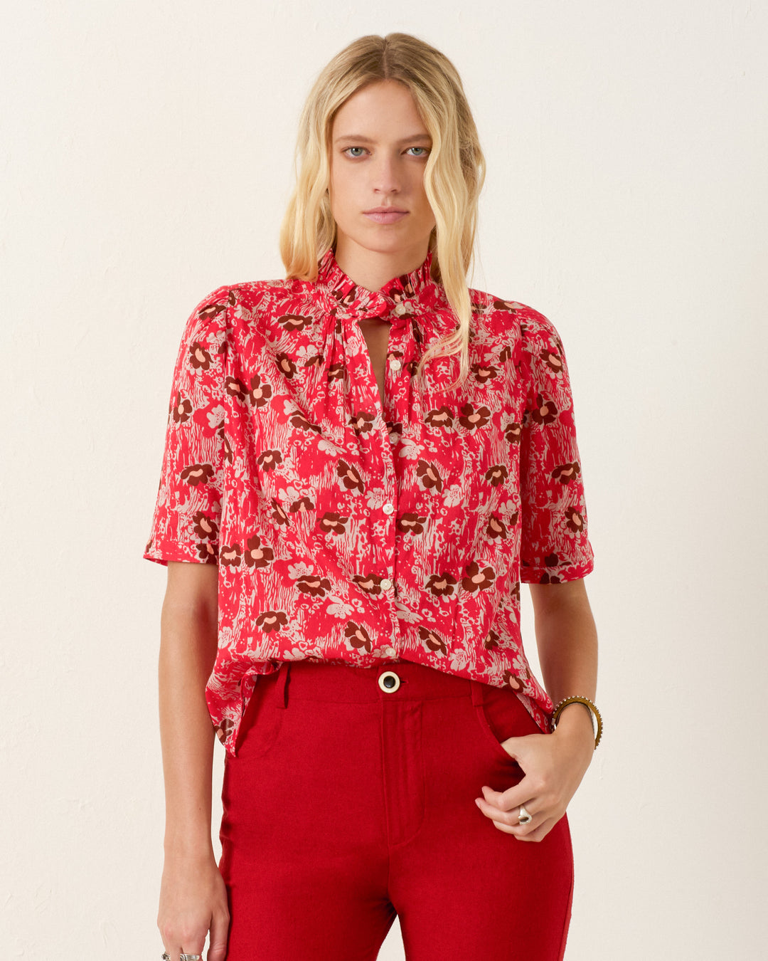 A woman wears the Winnie Rougewood Shirt, a red patterned blouse with a ruffled collar and short sleeves, paired with high-waisted red pants against a plain light background.