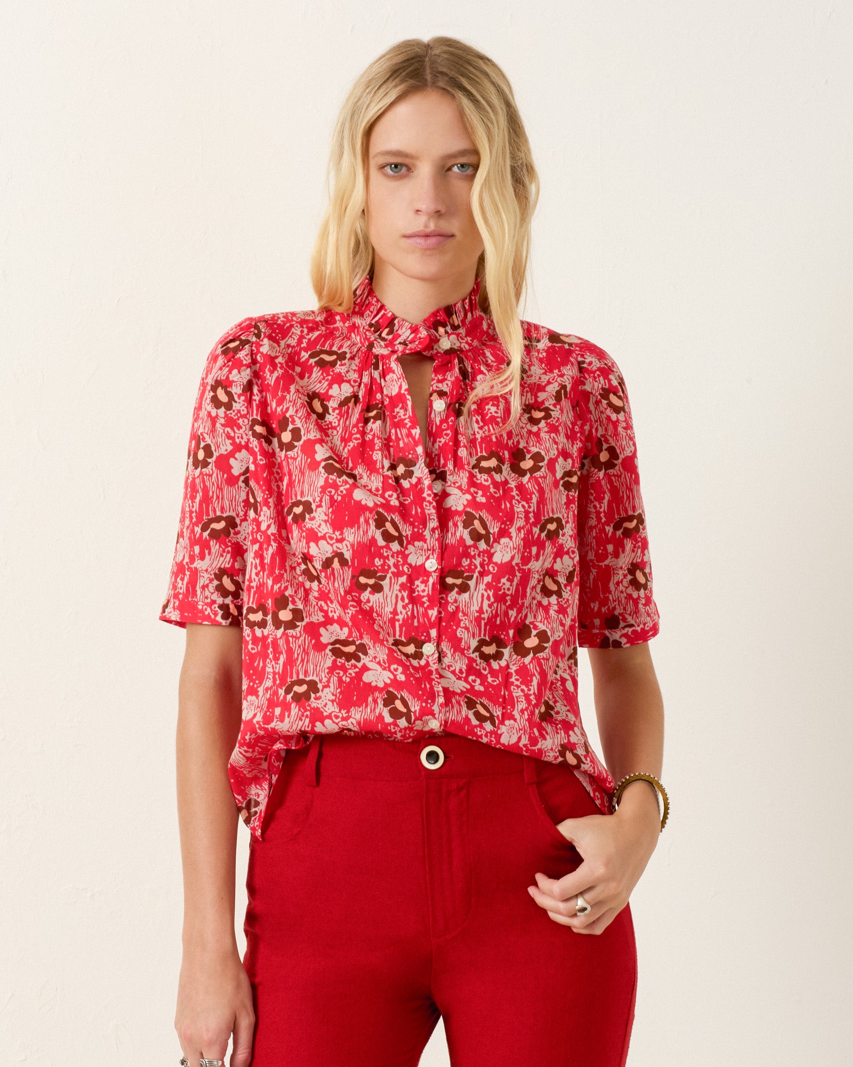 A woman wears the Winnie Rougewood Shirt, a red patterned blouse with a ruffled collar and short sleeves, paired with high-waisted red pants against a plain light background.